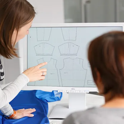 Dos personas miran patrones digitales de ropa deportiva en una pantalla de computadora, mientras una persona señala hacia la pantalla.