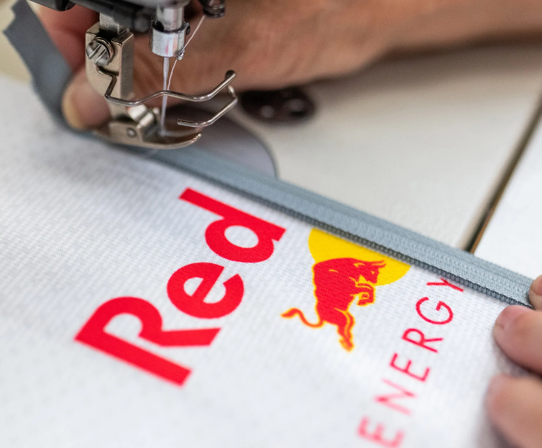 Una máquina de coser está cosiendo una cremallera gris en una prenda deportiva blanca con el logotipo de Red Bull.