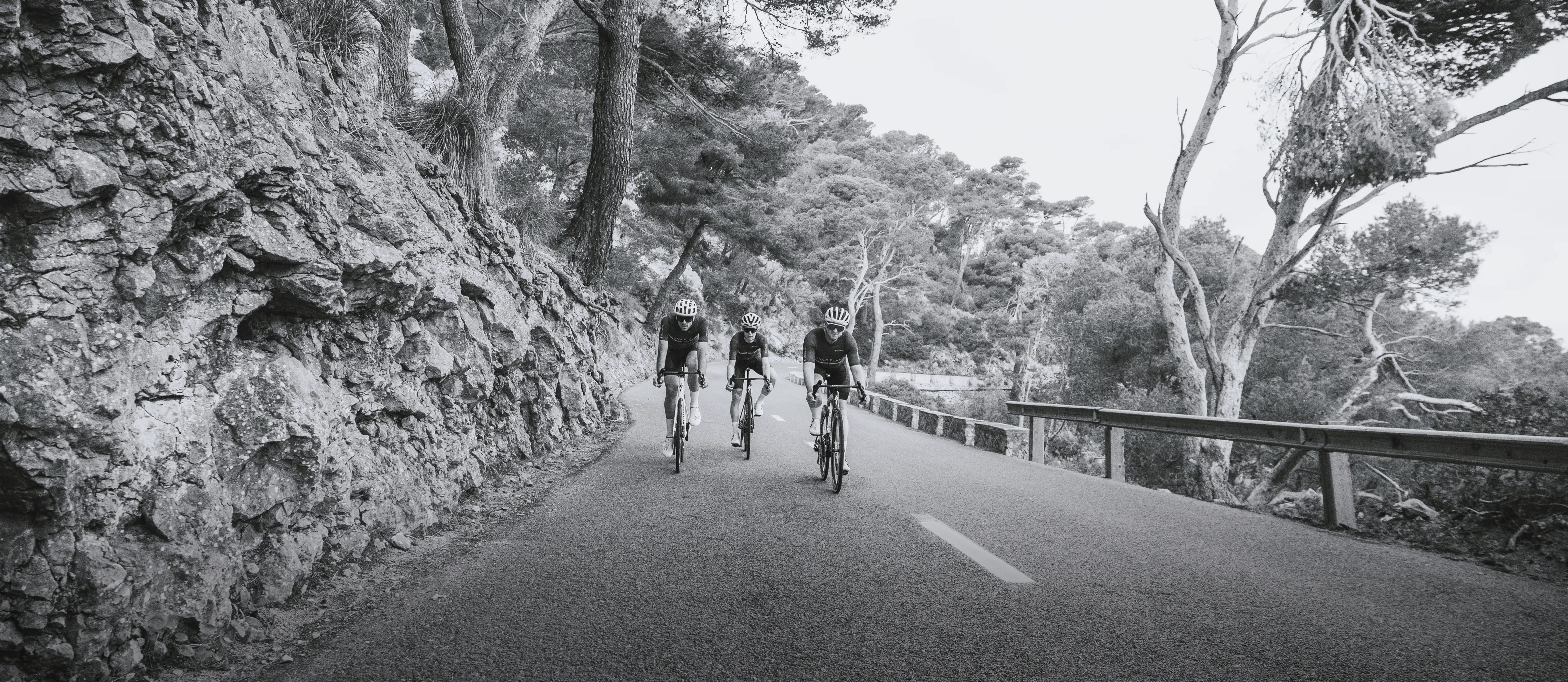 Tres ciclistas con ropa deportiva circulan por una carretera junto a un acantilado rocoso.