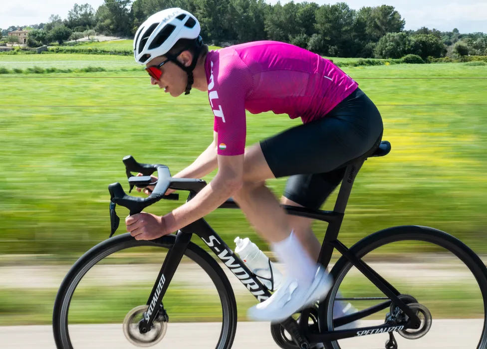 Un ciclista con un maillot rosa y pantalones cortos negros pedalea en una carretera a través de un paisaje verde, con desenfoque de movimiento que resalta la velocidad.