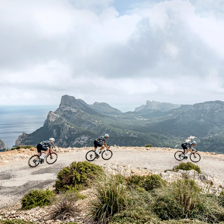 Tres ciclistas en ropa deportiva montan en una carretera sinuosa con un paisaje montañoso y el mar de fondo.