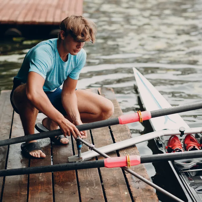 Un joven en una camiseta deportiva azul está reparando remos en un muelle junto al agua.