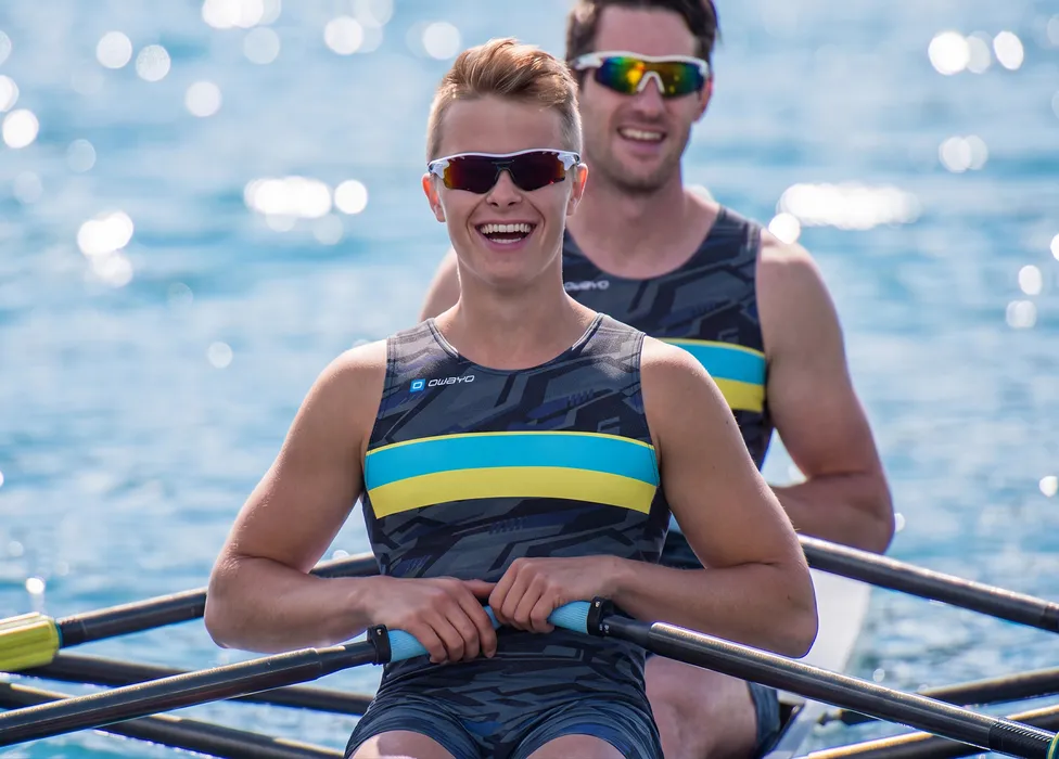 Dos remeros en trajes deportivos con patrones y franjas azules y amarillas, sonriendo en un cuerpo de agua.