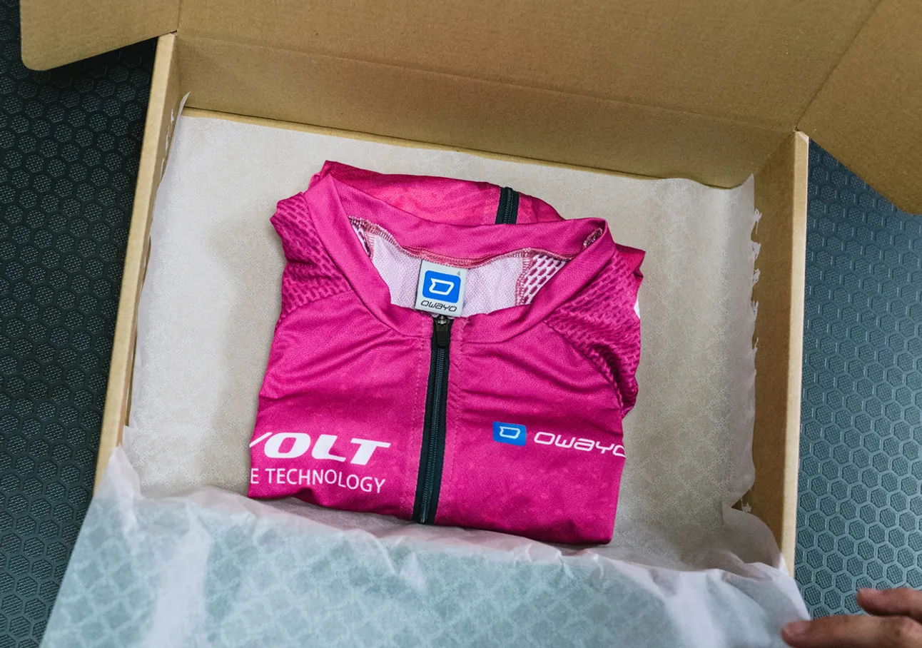 Una camiseta deportiva sin mangas de color rosa plegada con el logotipo de owayo está colocada en una caja de cartón sobre papel blanco.