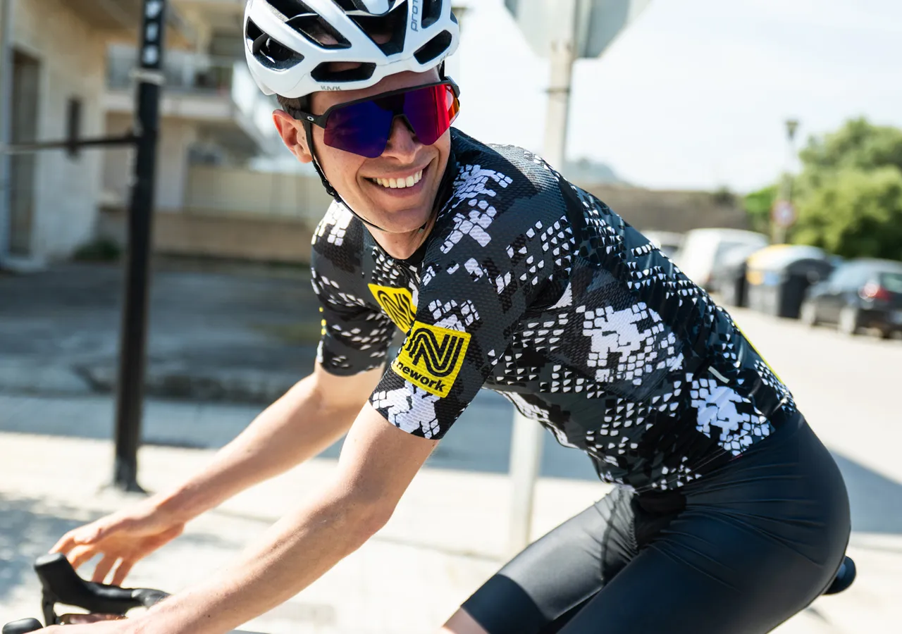 Un ciclista con una camiseta de ciclismo negra y blanca con un logotipo de patrocinador amarillo, pedaleando por una calle.
