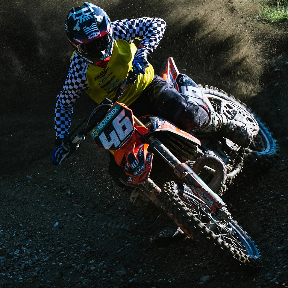 Un piloto de motocross con una chaqueta amarilla con mangas a cuadros negros y blancos y un casco conduce una motocicleta naranja en una pista polvorienta.