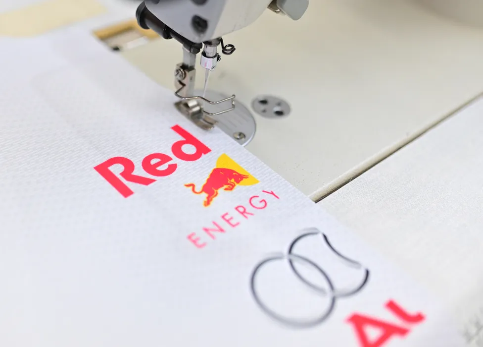 Una máquina de coser está cosiendo una prenda deportiva blanca con el logotipo rojo de Red Bull Energy y otro logotipo.