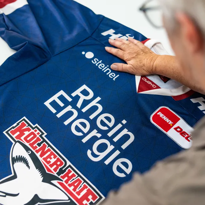 Una persona está revisando una camiseta deportiva azul personalizada de owayo con varios logotipos de patrocinadores, incluyendo 'Rhein Energie' y 'Kölner Haie'.