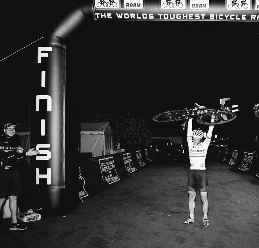 Un ciclista celebra cruzar la línea de meta de la 'Race Across America' levantando su bicicleta sobre su cabeza, mientras un hombre a su lado aplaude.