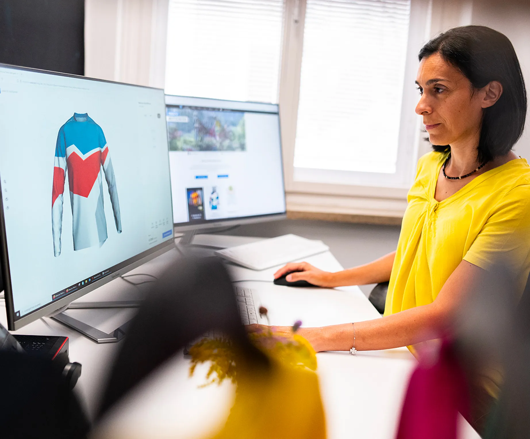 Una mujer con una camiseta amarilla está sentada en un escritorio y diseña en una pantalla de ordenador una camiseta deportiva personalizada con paneles azules, rojos y blancos.